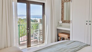 A bedroom with a balcony, a bed, a vanity with a mirror, and a view of the ocean. - Lavender Manor Guest Lodge (Hermanus)