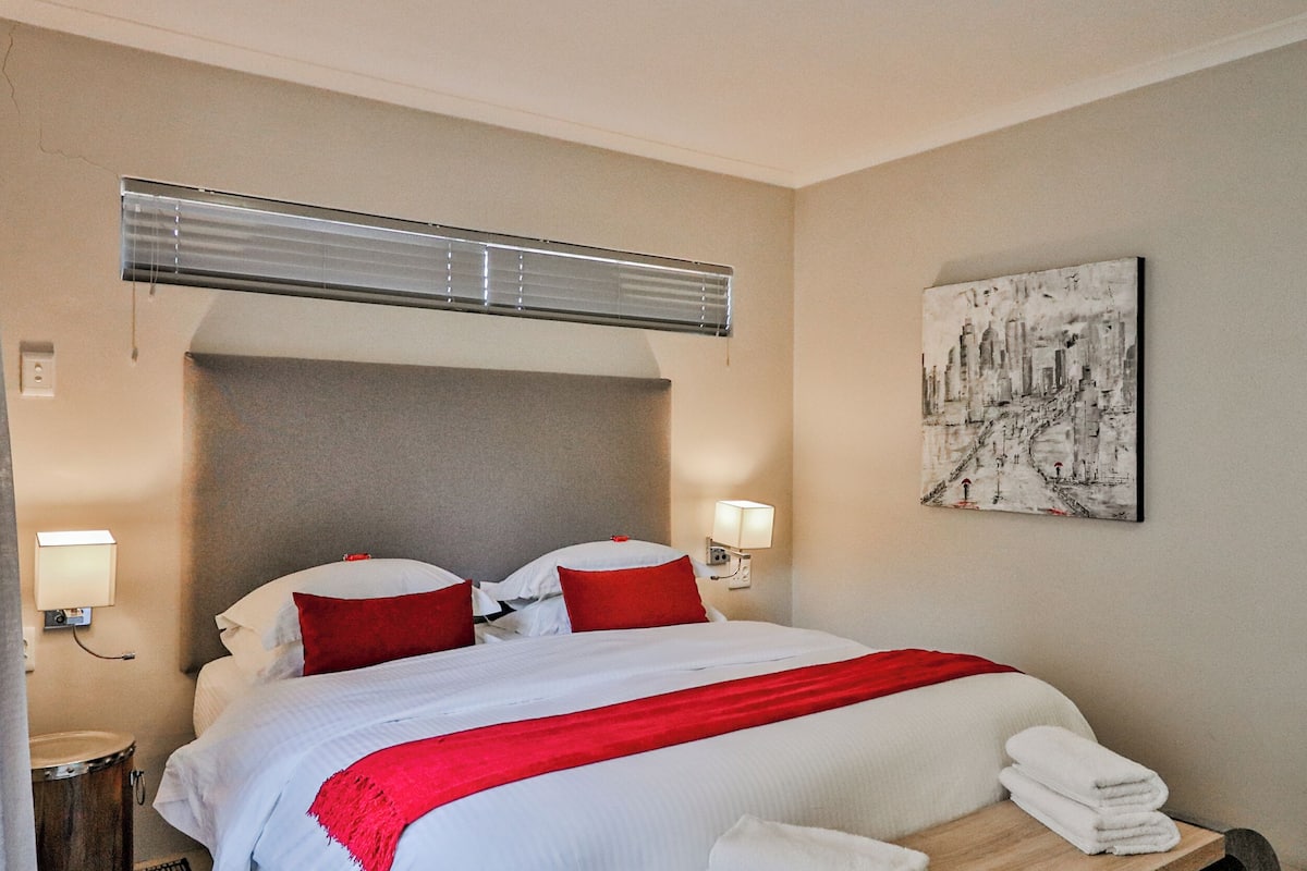Une chambre d’hôtel avec un lit, un plaid rouge, une tête de lit, un climatiseur fixé au mur, un tableau et une table de chevet avec des serviettes.