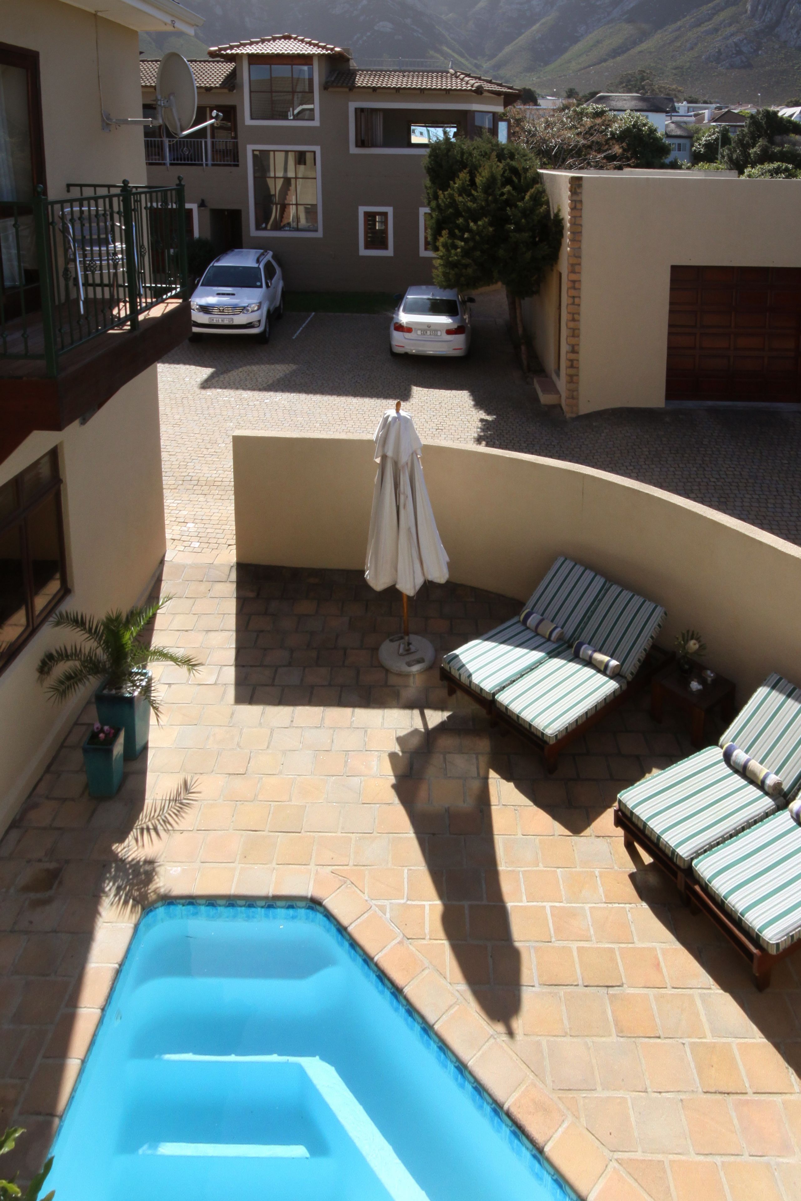 Ein Poolbereich mit Liegestühlen, einem kleinen Tisch mit Sonnenschirm und Blick auf eine Wohnstraße mit Autos und Häusern.