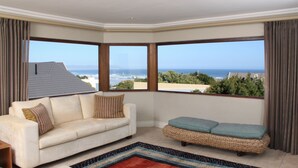 A room with a white sofa, a woven bench, and a large window offering a view of the ocean. - Lavender Manor Guest Lodge (Hermanus)