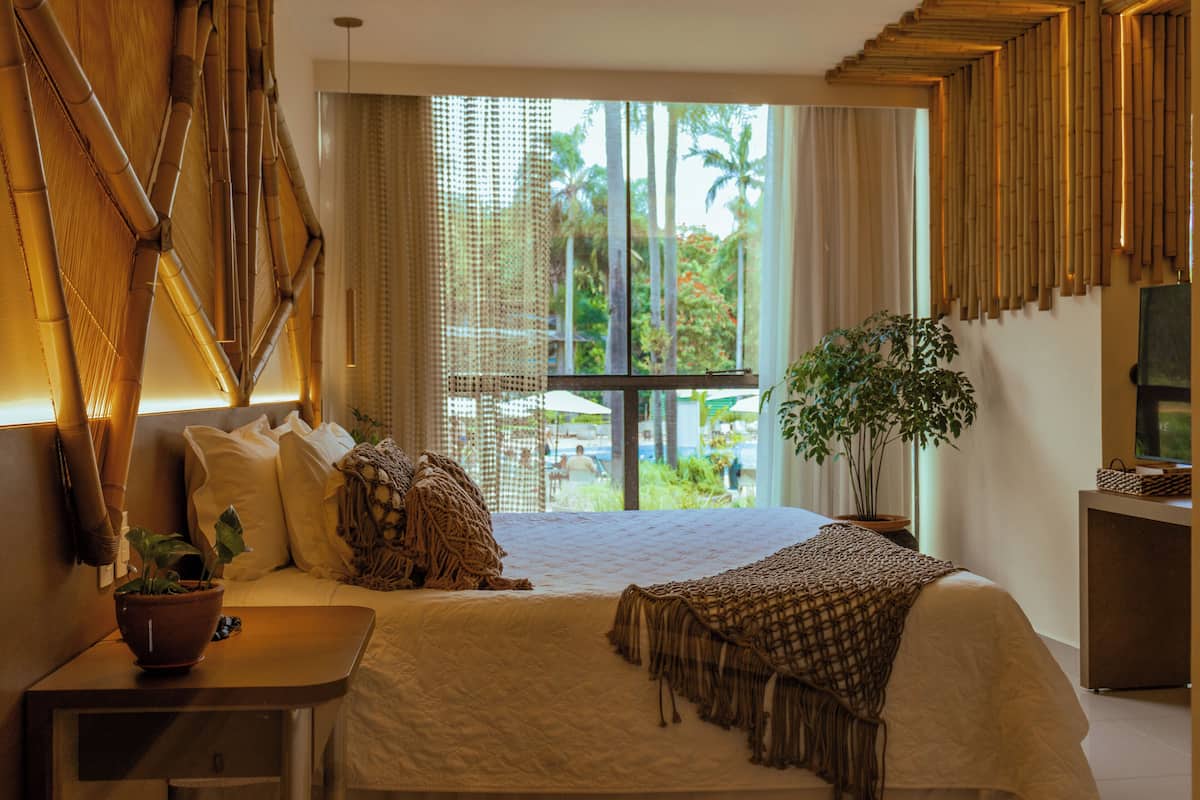 Ein Hotelzimmer mit einem großen Bett, einem Nachttisch mit Pflanze, Blick auf Palmen und einer Wandgestaltung mit Bambus.
