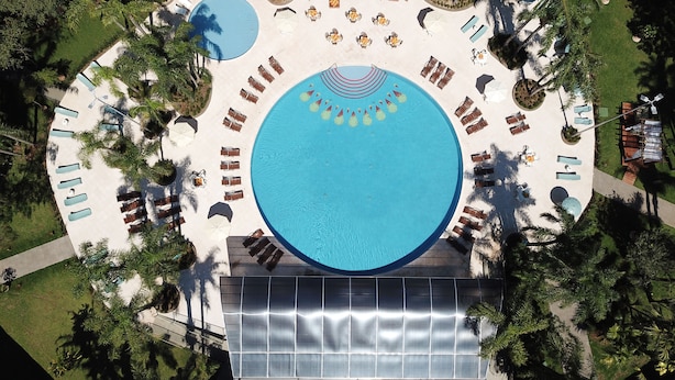 An aerial view of a resort with a large circular pool surrounded by sun loungers and umbrellas, palm trees, and a covered seating area.