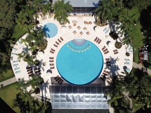 Vista aérea de un complejo turístico con una amplia piscina circular rodeada de tumbonas y sombrillas, palmeras y una zona de estar cubierta.