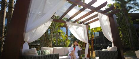 Un espacio al aire libre cubierto con sofá y sillas, rodeado de follaje y una pérgola de madera.