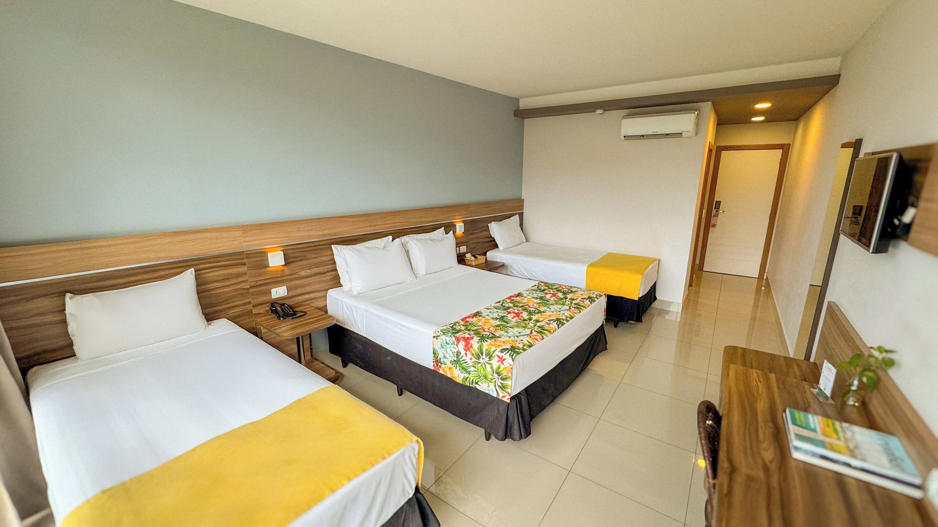 Ein Hotelzimmer mit zwei Betten, einem Holz-Kopfteil, einem Nachttisch mit Telefon, einem Schreibtisch mit Pflanze und einem an der Wand befestigten Fernseher.