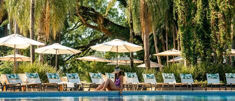 A poolside area with lounge chairs and umbrellas, surrounded by lush greenery and tall trees.