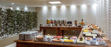 Montaje de bufé con diversos alimentos y bebidas, una mesa de madera y una pared decorativa con un diseño ondulado.