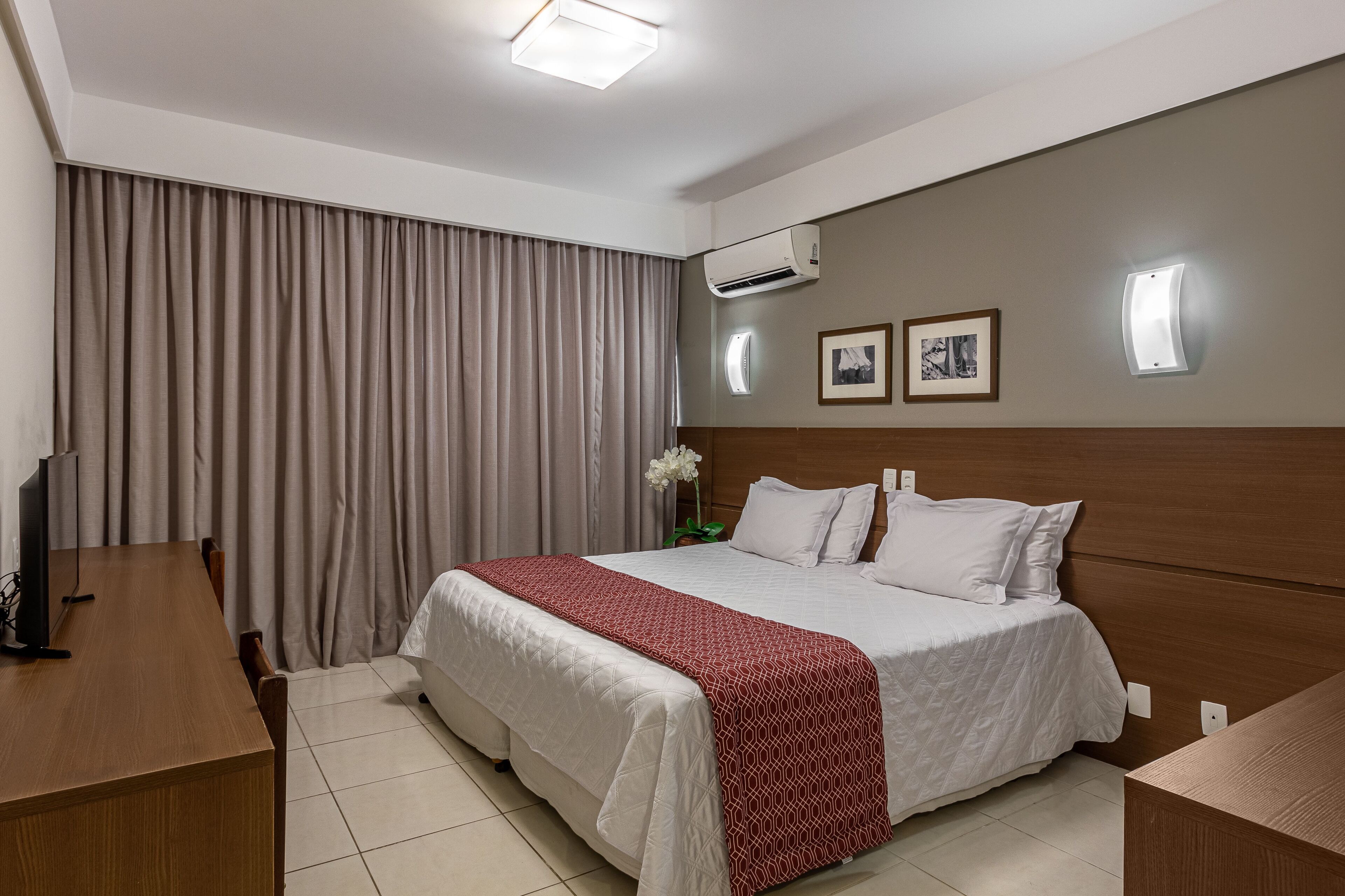 A hotel room with a bed, a desk, a chair, an air conditioning unit, and a wall-mounted light.