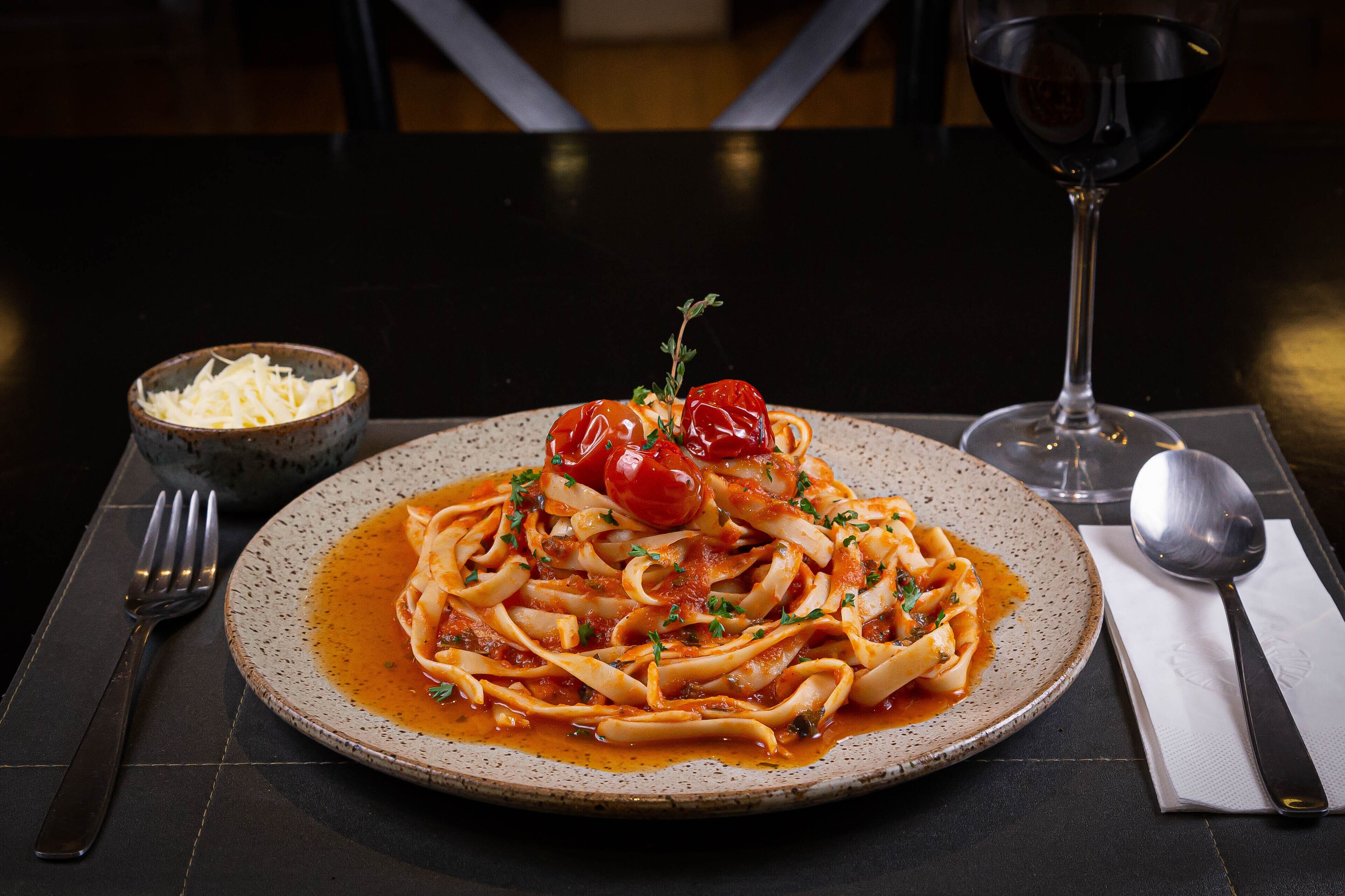 Een bord pasta met kerstomaatjes en kruiden, een glas rode wijn en een schaal geraspte kaas op een tafel.