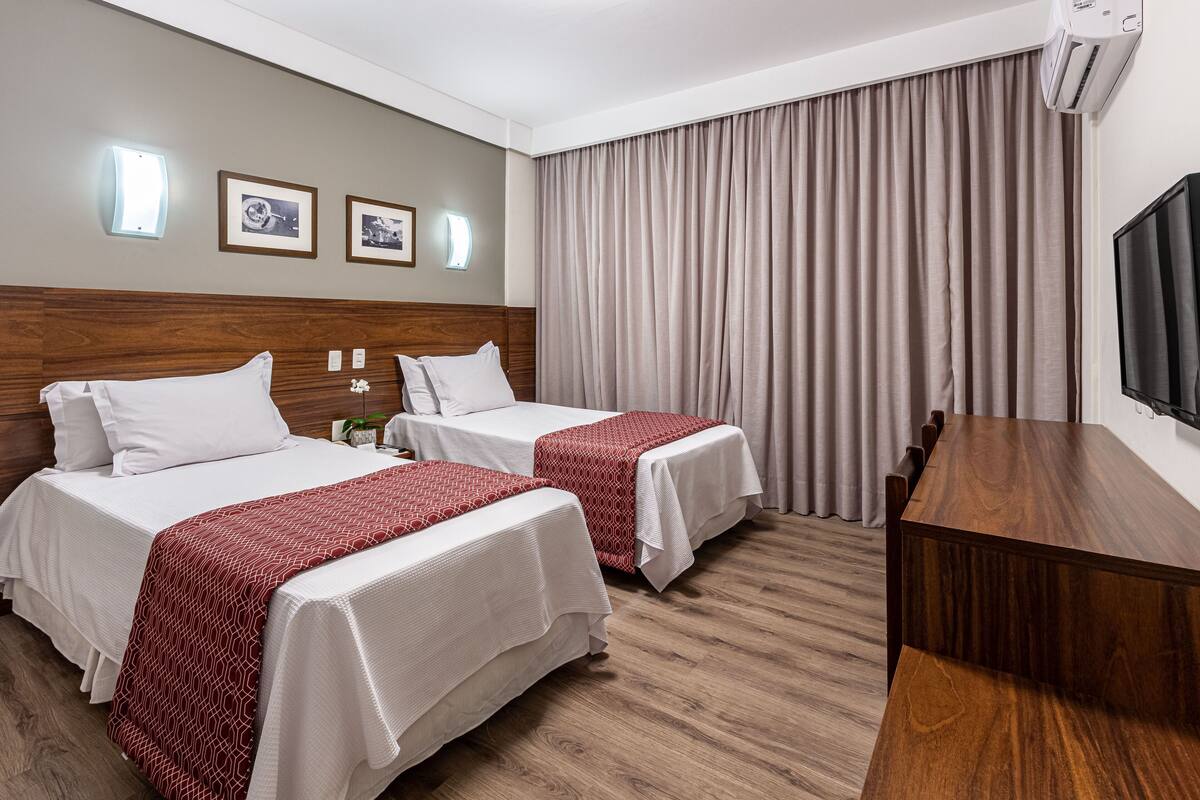 A hotel room with two beds, a wooden desk, a TV, and a large window with curtains.