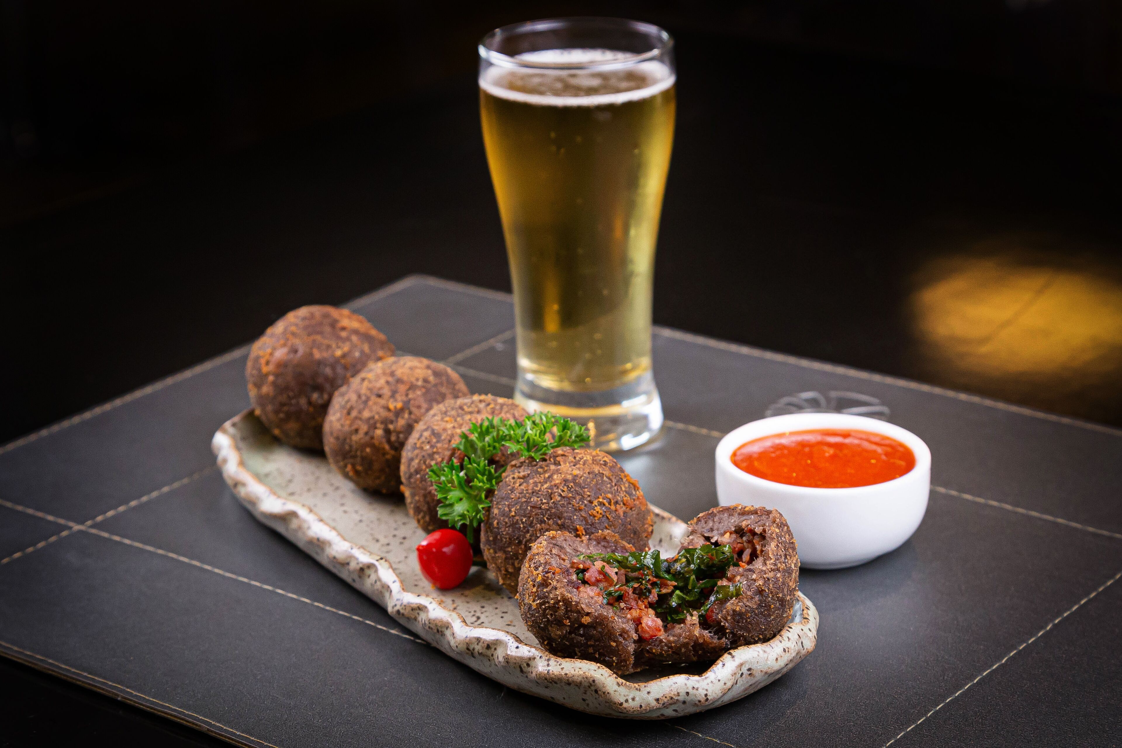 Ein Teller Hackbällchen mit roter Sauce, ein Glas Bier und eine kleine Schale Soße auf einem dunklen Tisch.