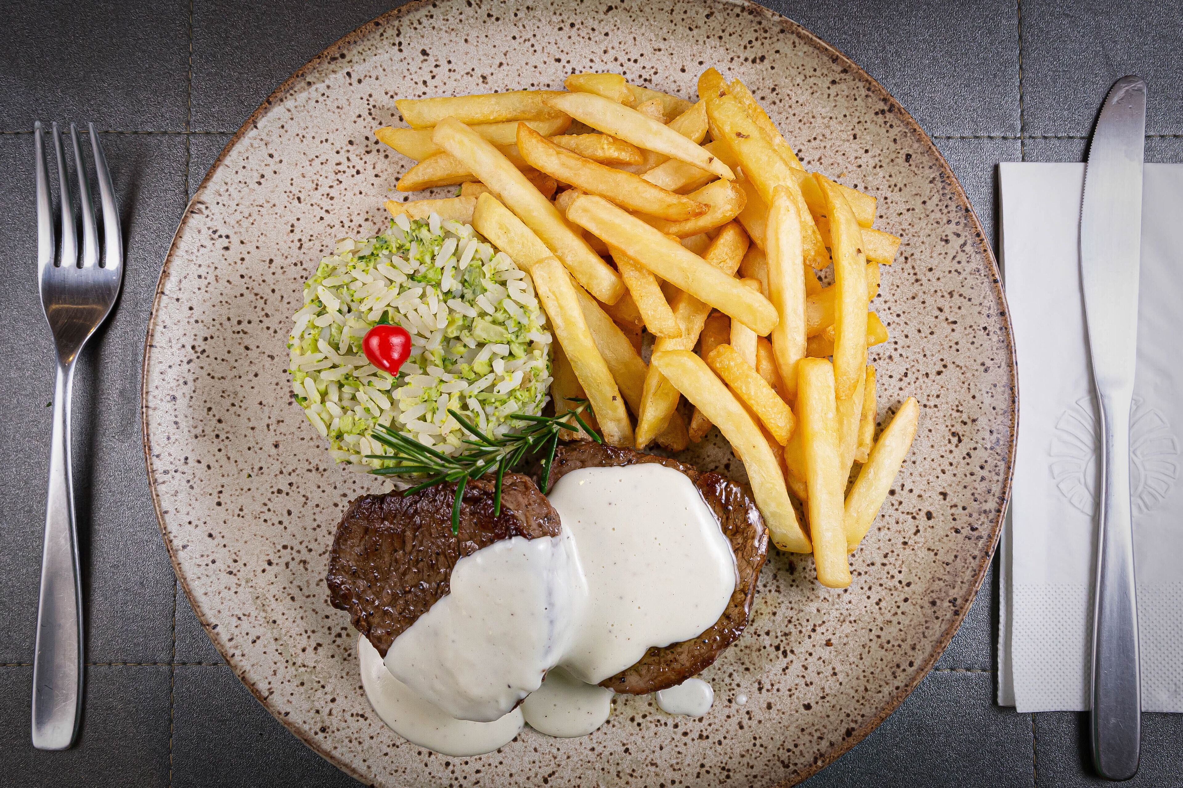 Un piatto con una bistecca ricoperta di salsa, patatine fritte e un contorno di riso con una ciliegia.