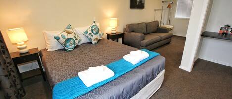 A hotel room with a bed, a sofa, a nightstand with a lamp, and a window with blinds.
