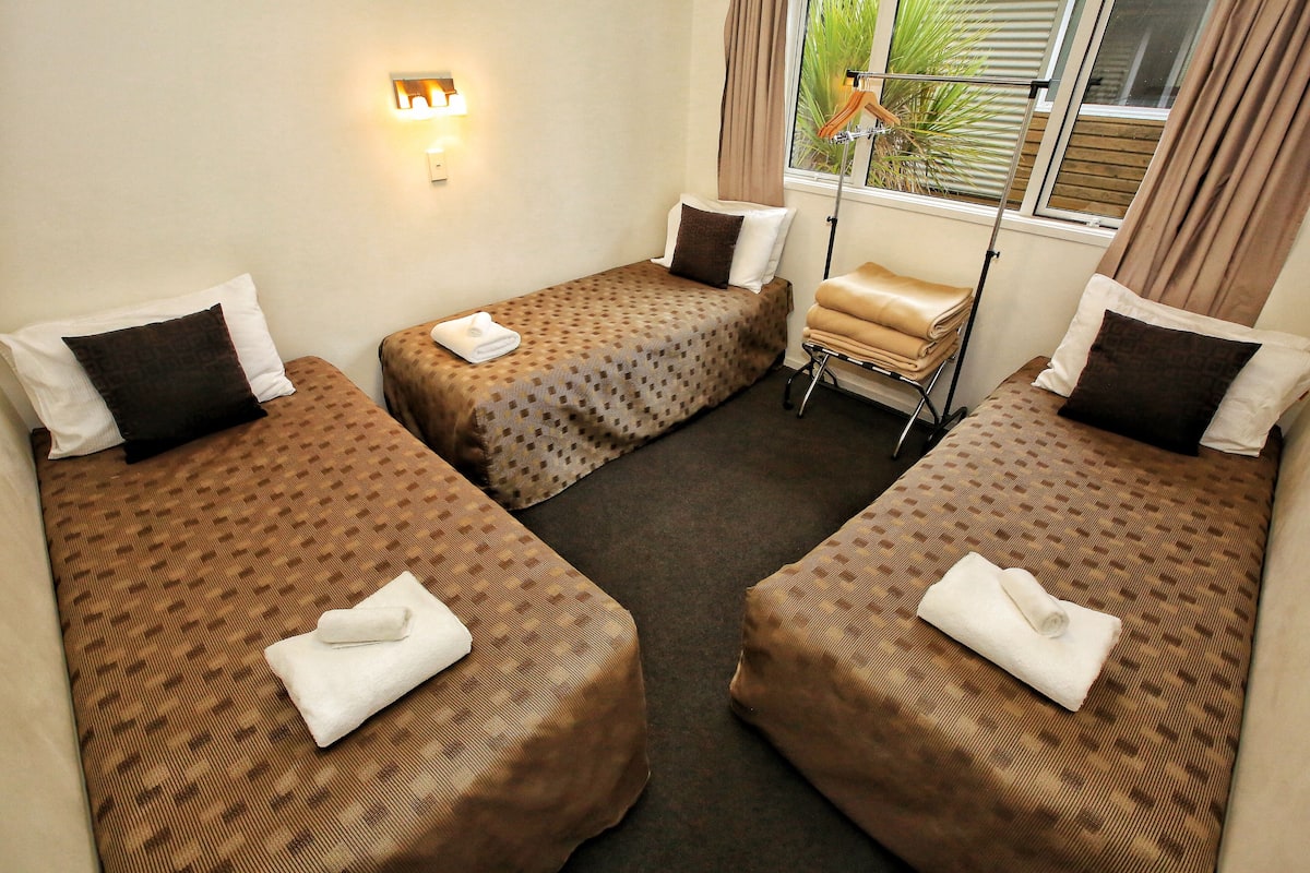 Habitación de hotel con tres camas individuales, cada una con una colcha marrón estampada y almohadas blancas. Hay una mesita con una silla y una lámpara cerca de la ventana.