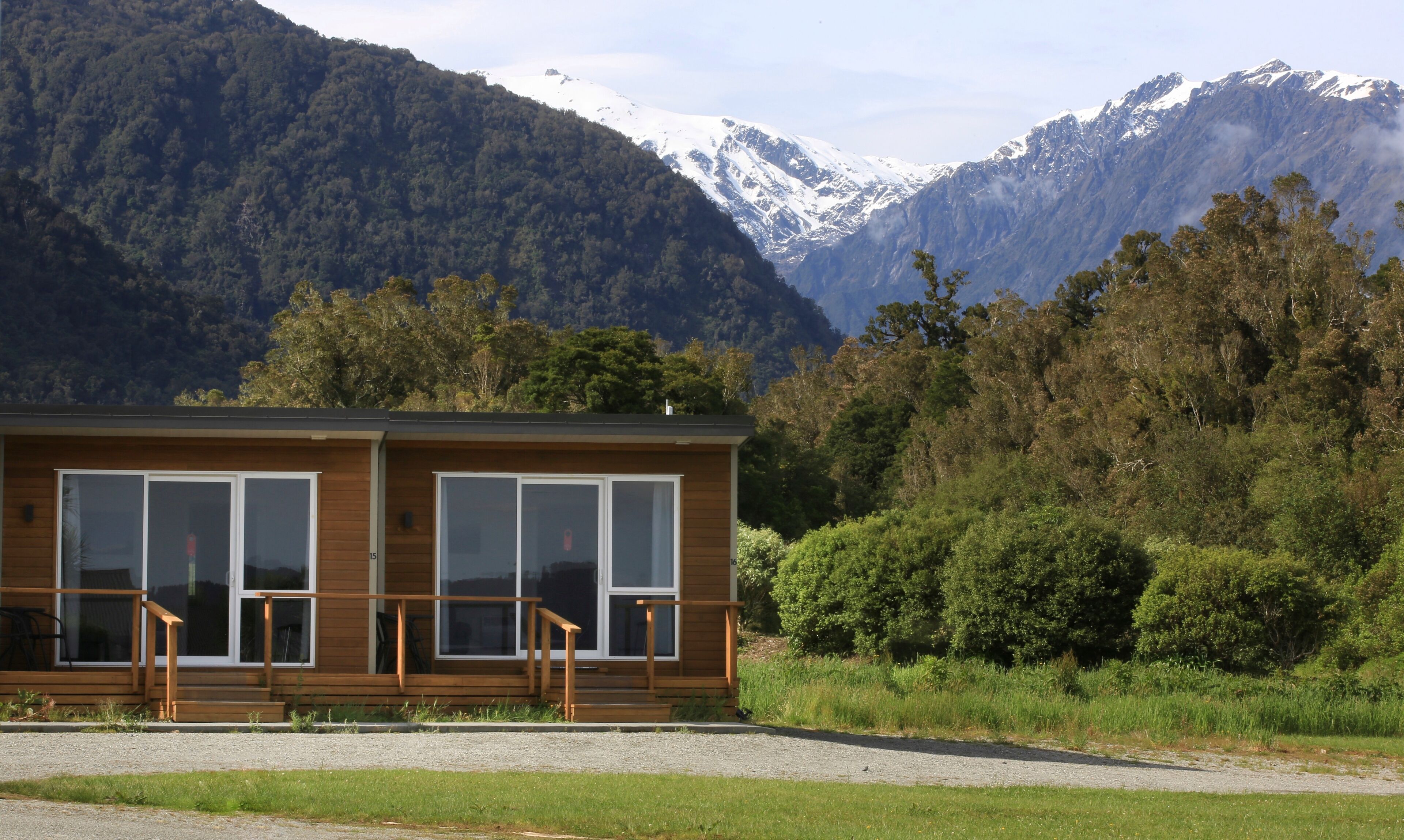 Une cabane en bois dotée de grandes fenêtres et d’un porche, entourée d’une végétation luxuriante et de montagnes.