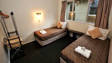 A small hotel room with two single beds, a chair, a bedside table, and a window with curtains.