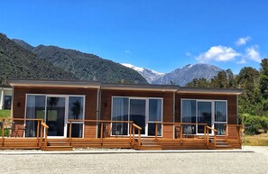 A modern wooden cabin with large sliding glass doors, a wooden deck, and a mountain backdrop. - Franz Alpine Retreat (Franz Josef Glacier)