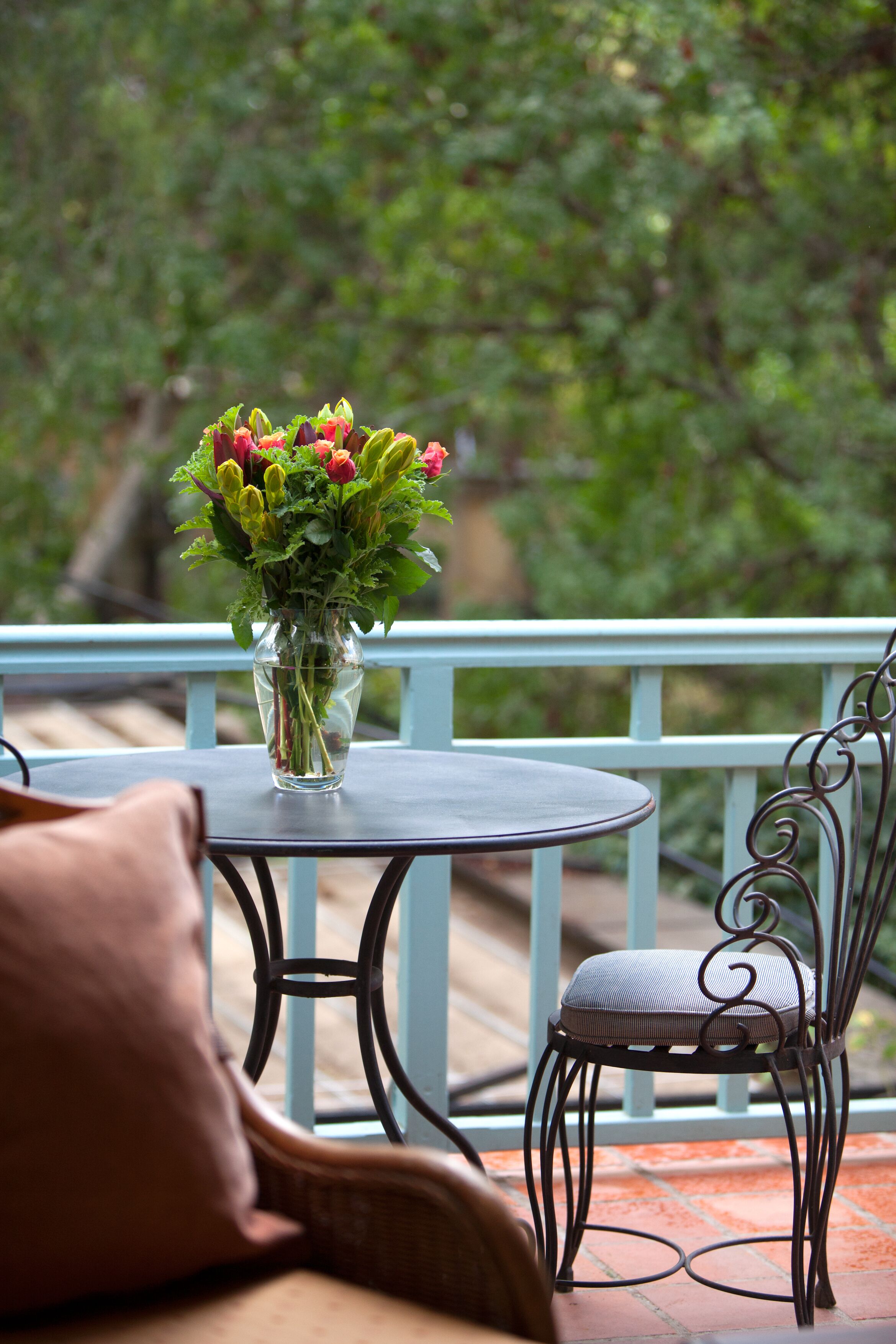 Een ronde tafel met een glazen vaas gevuld met bloemen, een rieten stoel en een blauwe balustrade.