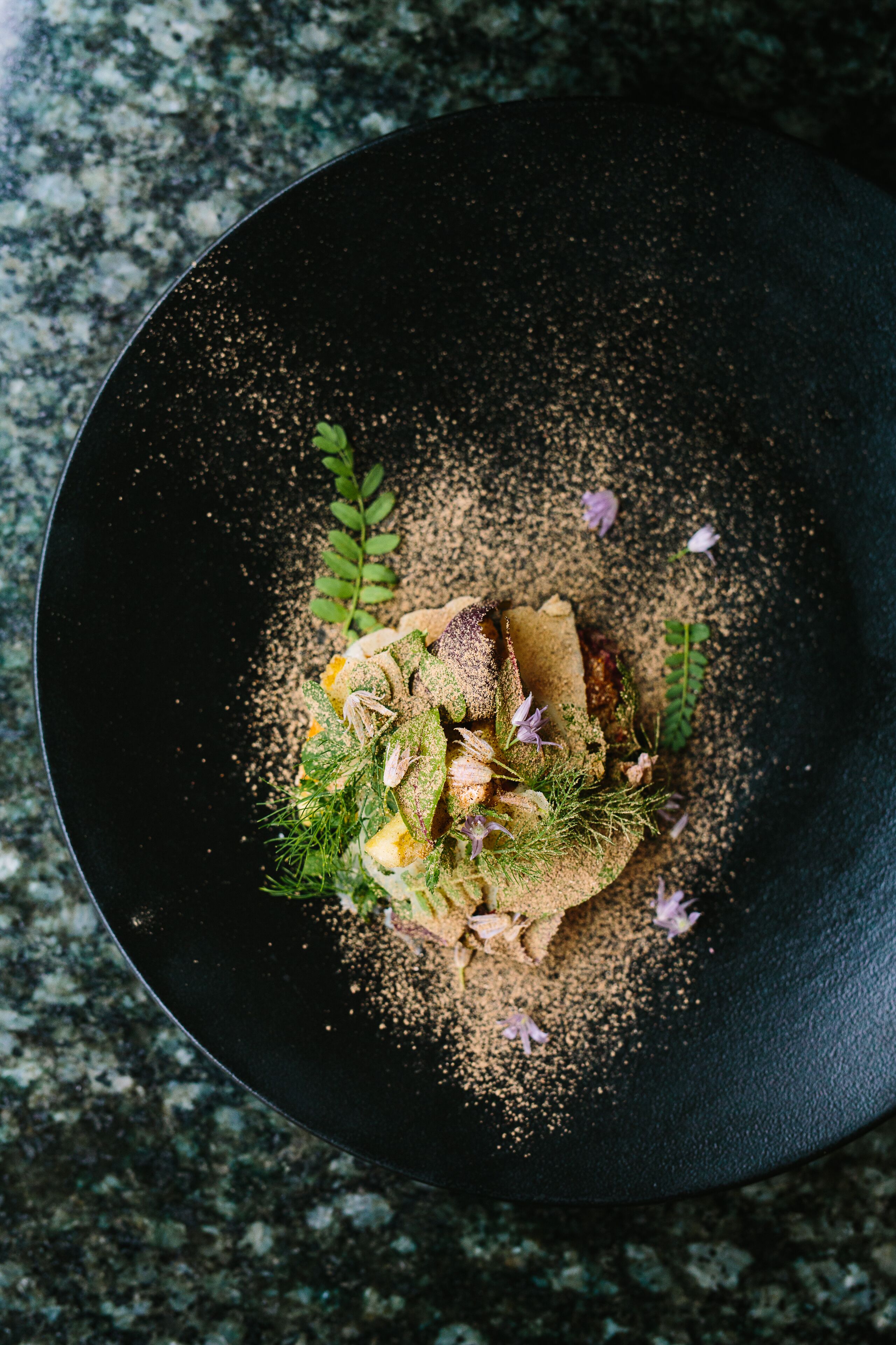 A black plate with a dish garnished with herbs and flowers on a granite countertop.