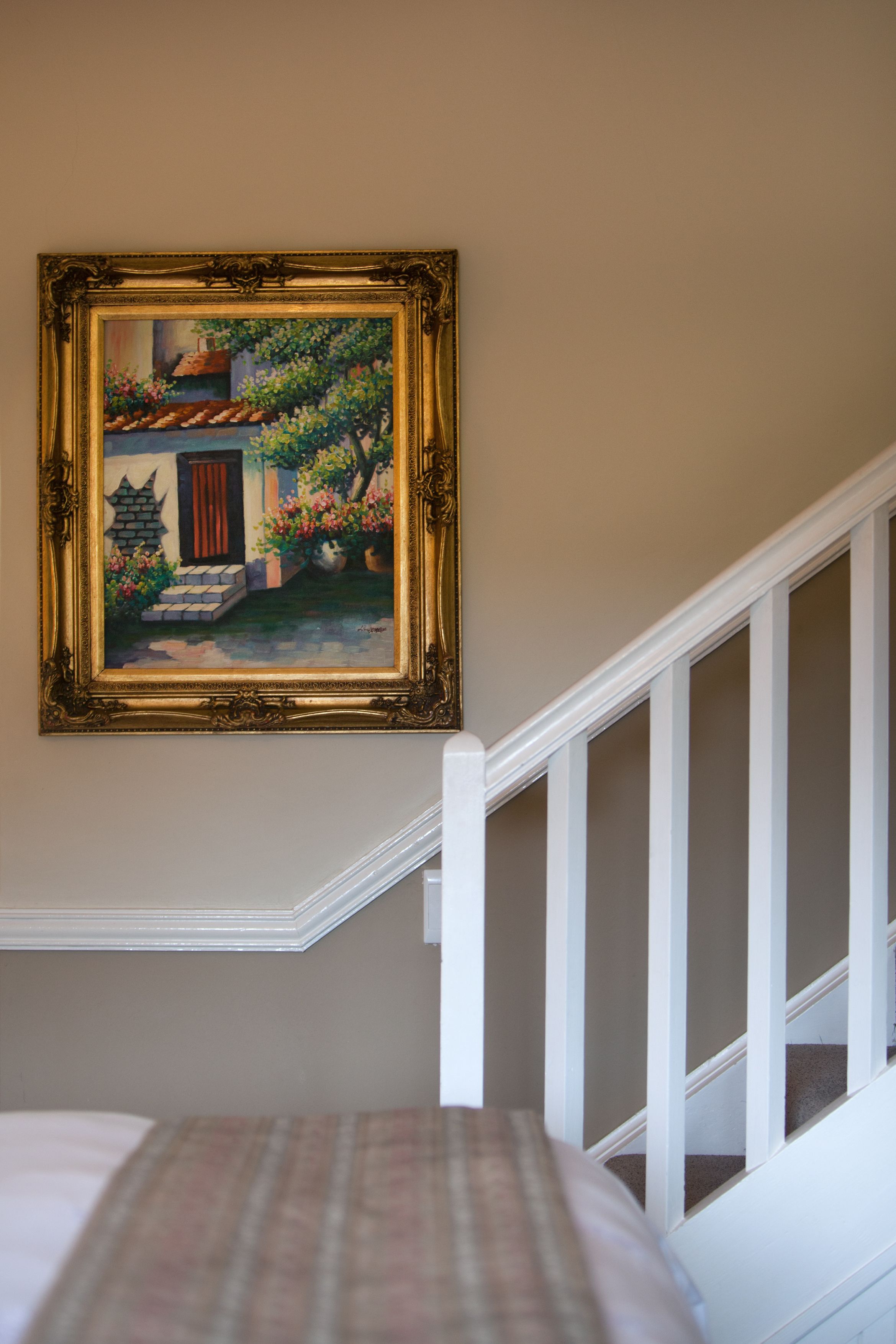 A bedroom with a bed, a painting of a house, and a white staircase.