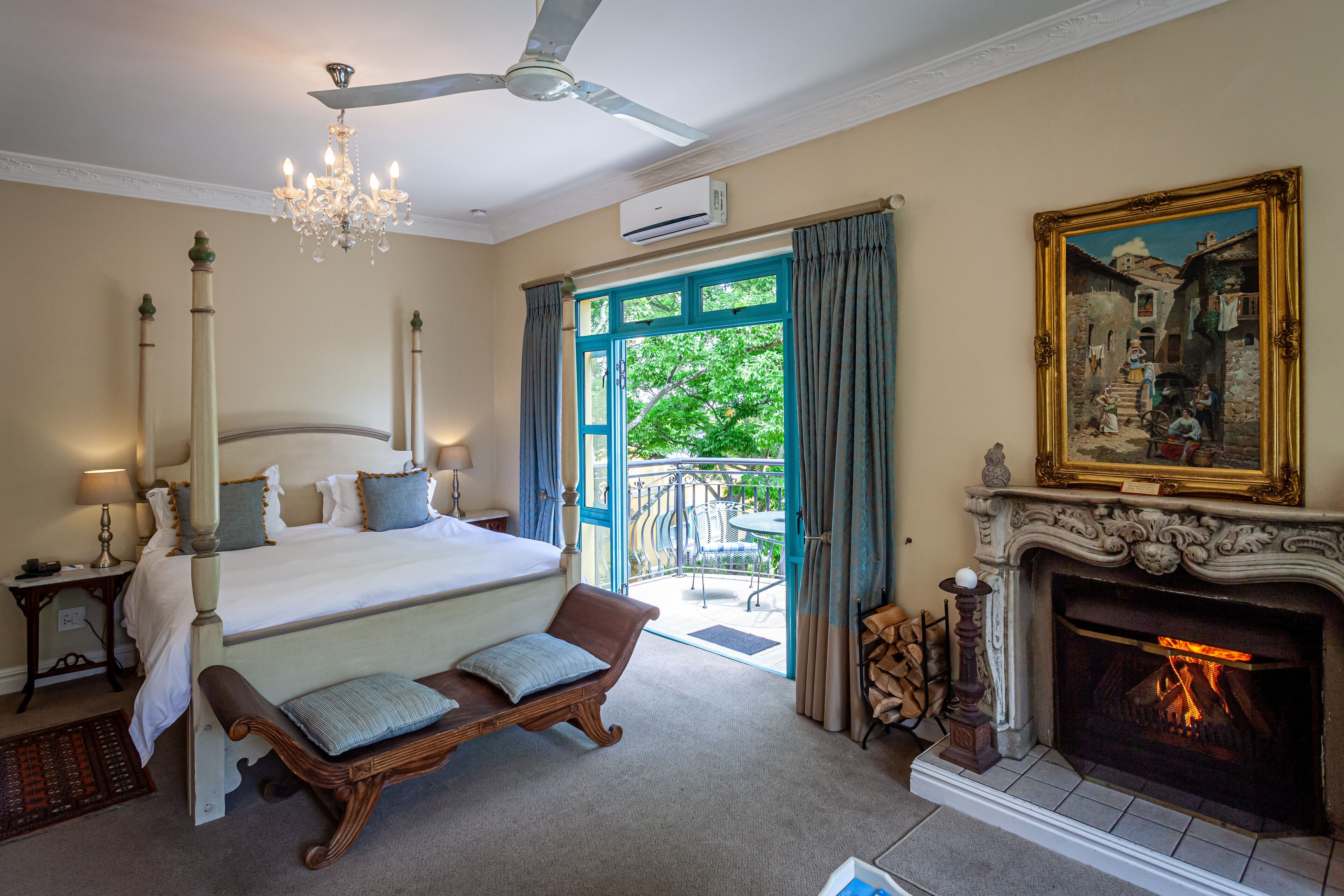 A bedroom with a large bed, a chandelier, a fireplace, and a balcony with a view of greenery.