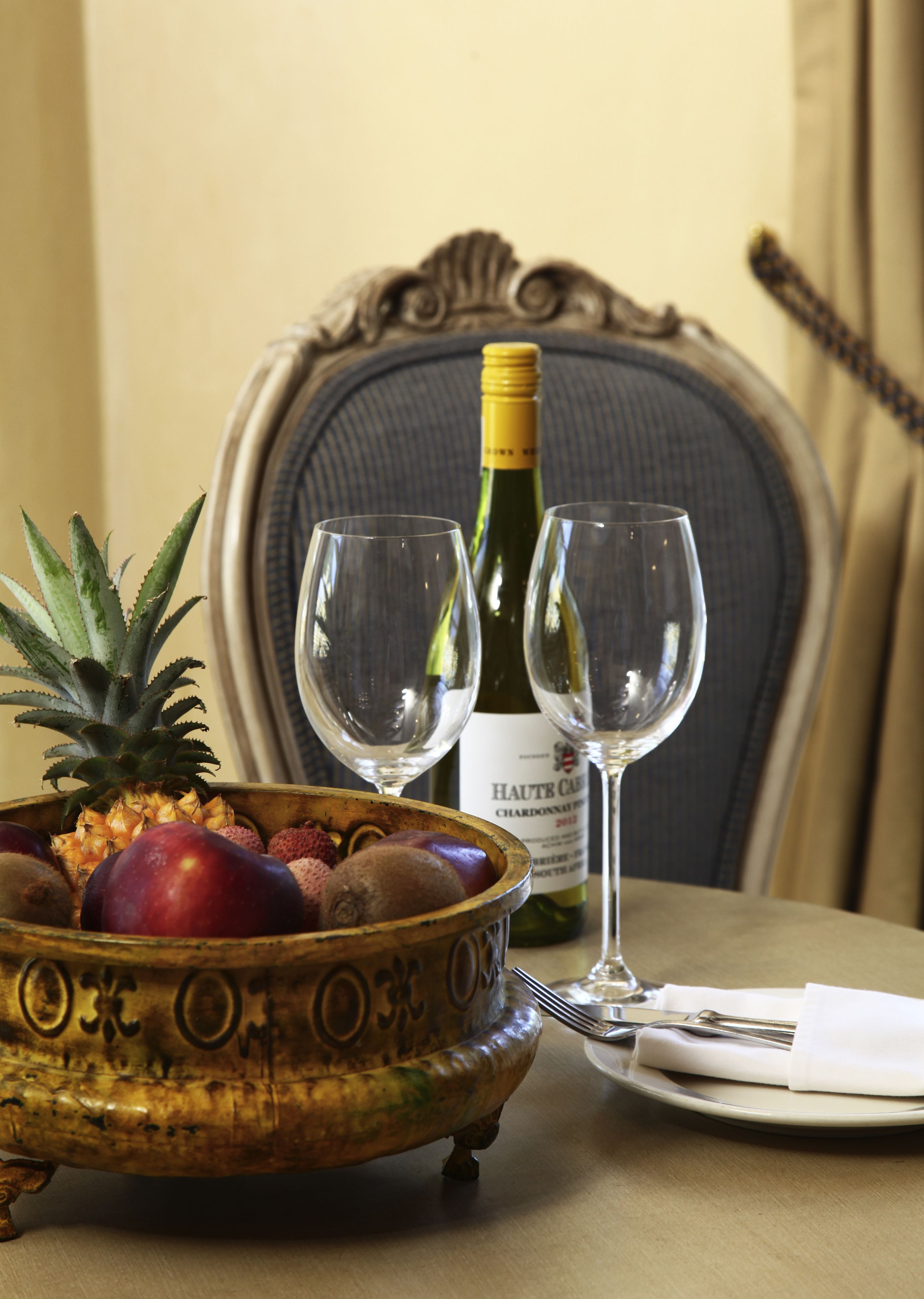 A dining table with a fruit bowl, two wine glasses, a bottle of wine, and a chair.