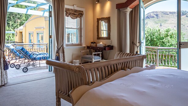 A bedroom with a large bed, a chandelier, and a view of a balcony with a chair and a stroller. - Franschhoek Country House and Villas (Franschhoek)