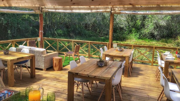 Une terrasse extérieure en bois, agrémentée de tables et de chaises, entourée d’une balustrade en bois et offrant une vue sur un paysage verdoyant luxuriant.