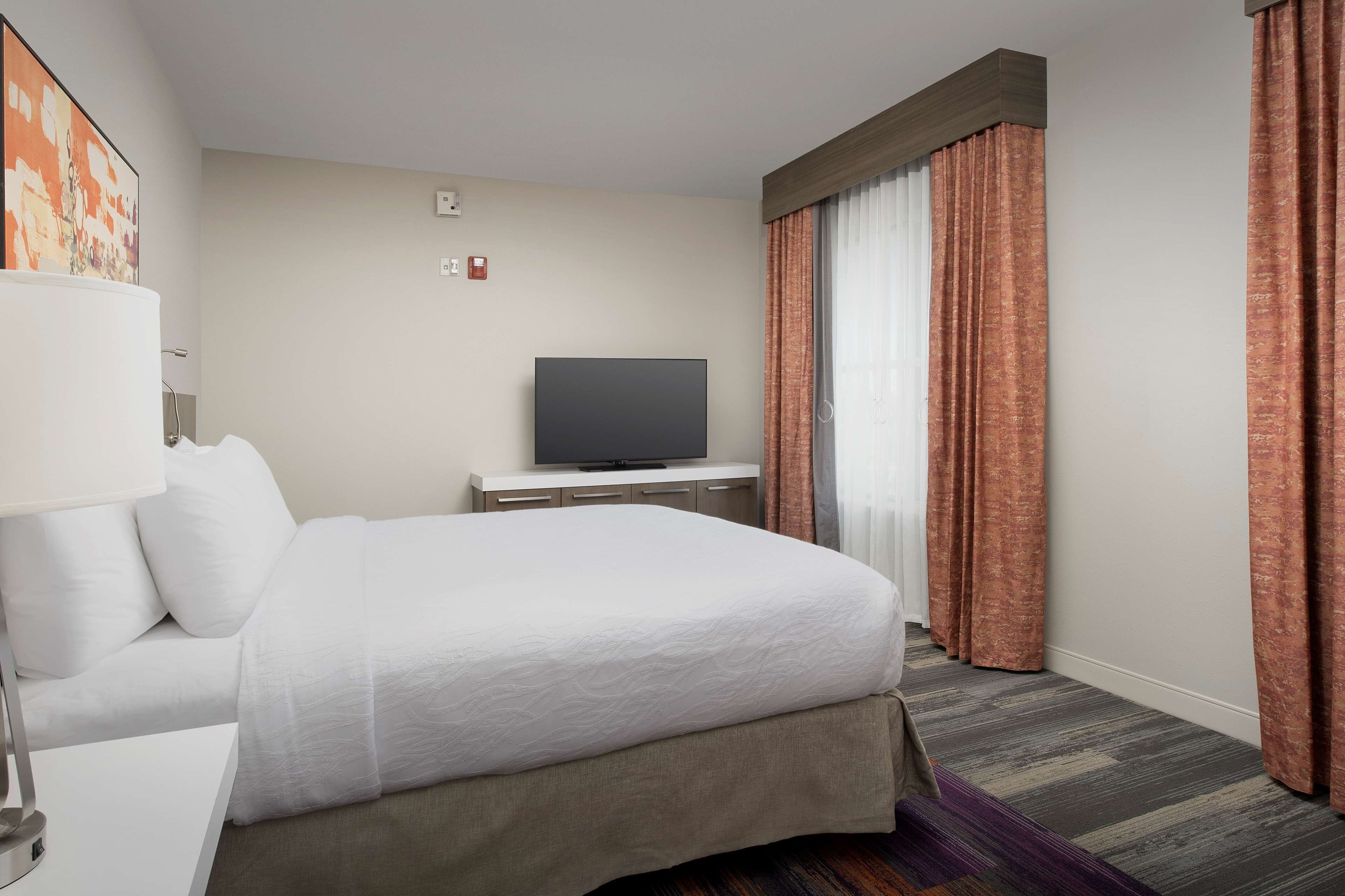 A hotel room with a bed, a flat-screen TV, a desk with a lamp, and a window with curtains.