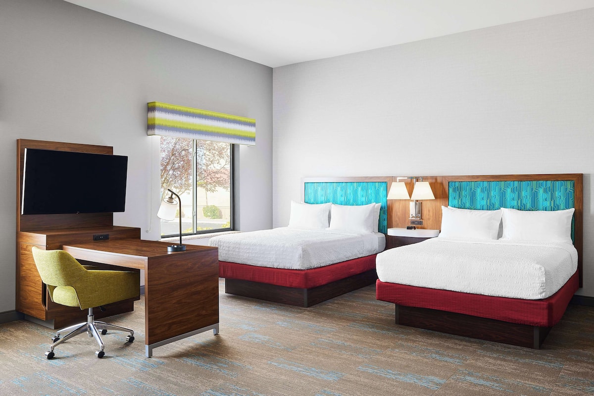 A hotel room with two beds, a desk with a chair, a TV, and a window with a view of trees.