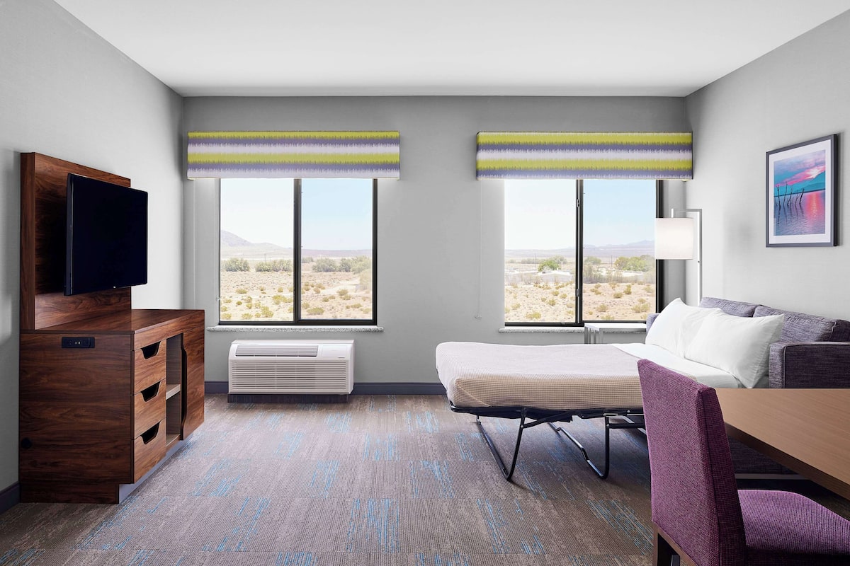 A hotel room with a wooden dresser, a flat-screen TV, a bed, a desk, a chair, and two windows with striped curtains.