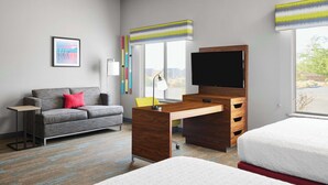 A hotel room with a grey sofa, a wooden desk, a flat-screen TV, a bed, and a window with a view of the outdoors. - Hampton Inn & Suites Ridgecrest (Ridgecrest)
