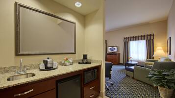 A hotel room with a kitchenette, a large mirror, a flat-screen TV, a sofa, a coffee maker, and a potted plant.
