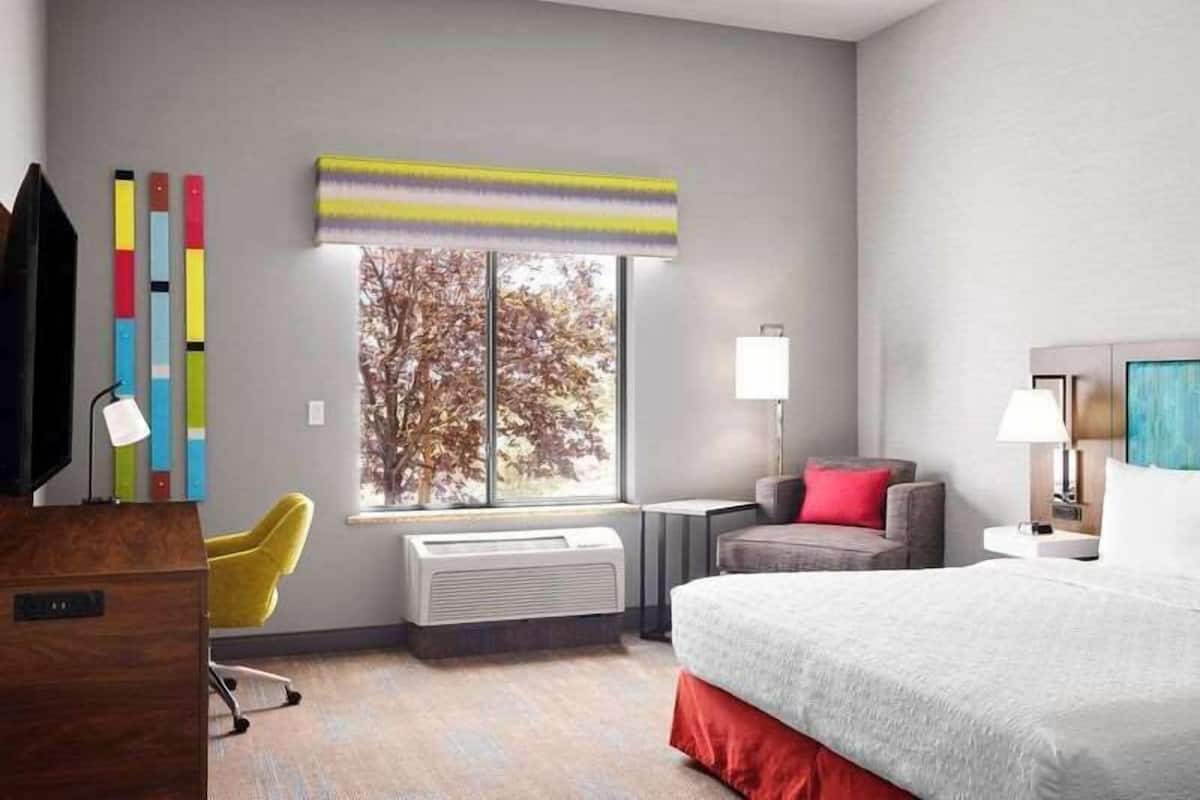A modern hotel room with a bed, a desk, a chair, a window with a view of trees, and a wall with colorful decor.