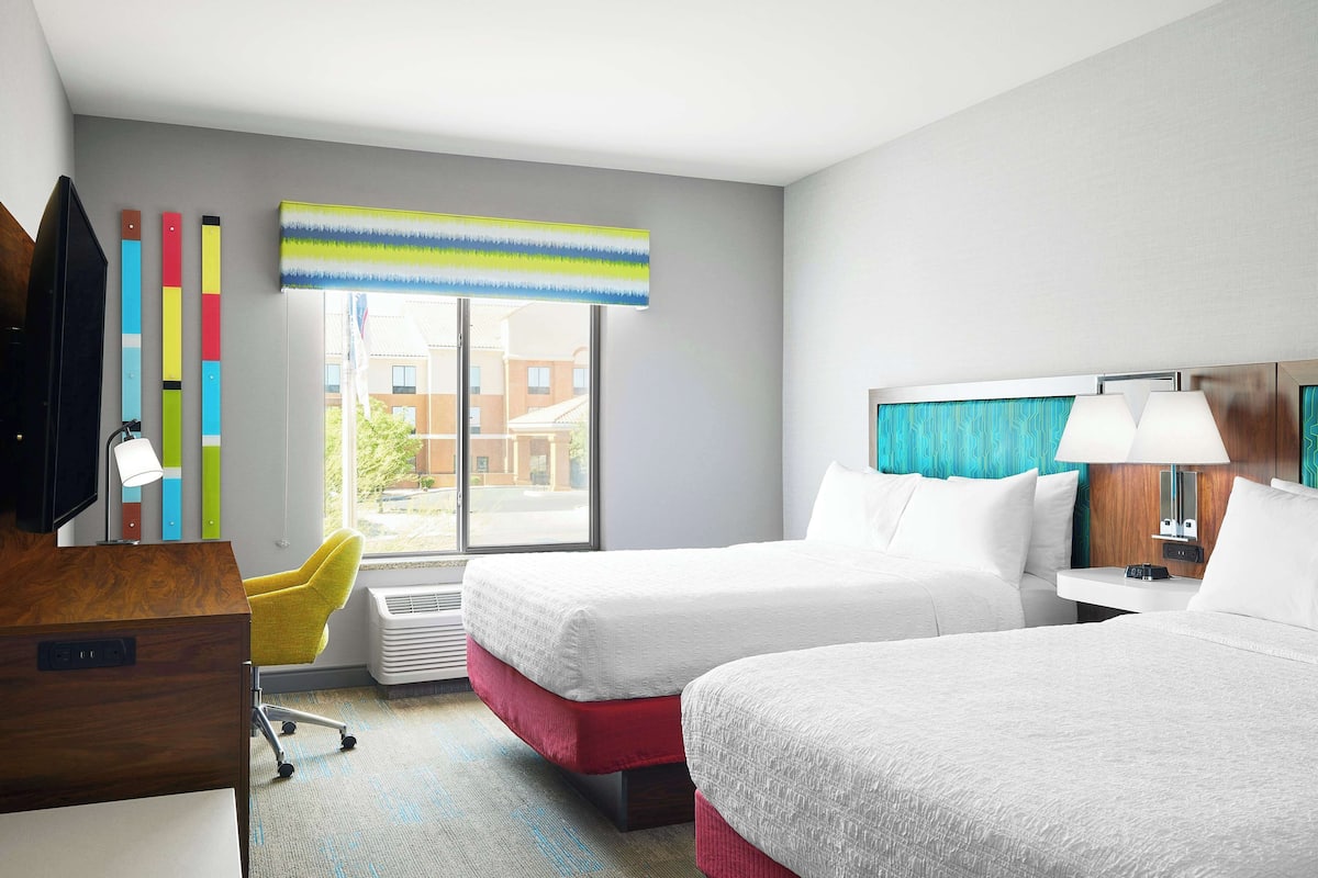 A hotel room with two beds, a desk, a chair, a window with blinds, and a view of buildings outside.