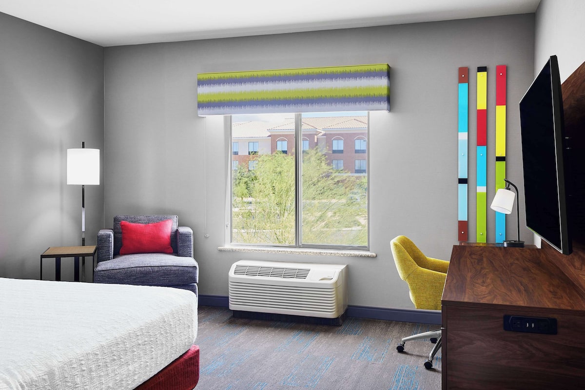 A hotel room with a bed, a desk, a chair, a window with a view, and an air conditioner.
