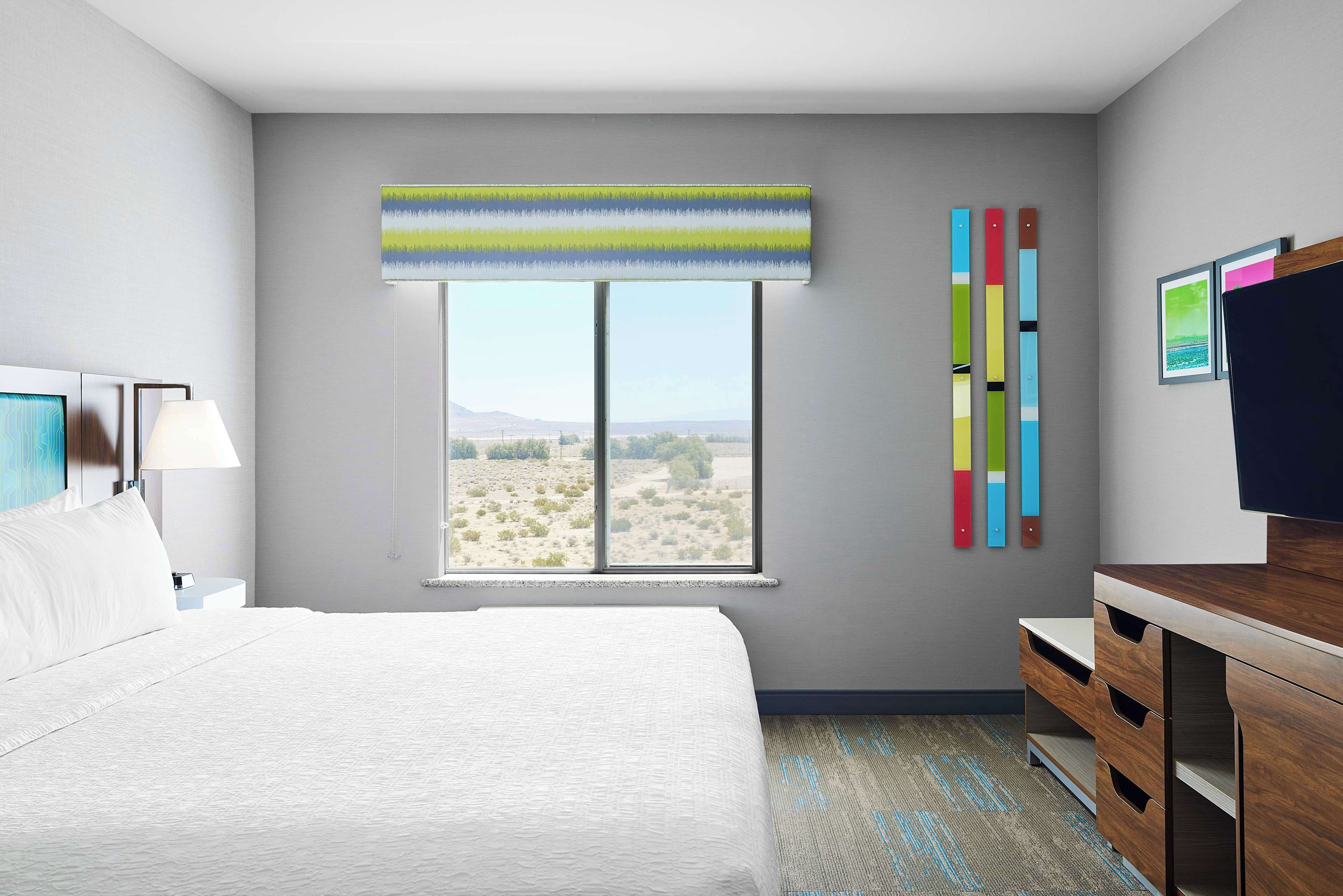 Habitación de hotel con una cama grande, un escritorio, una silla, un ventanal con vistas al desierto y una obra de arte colorida en la pared.