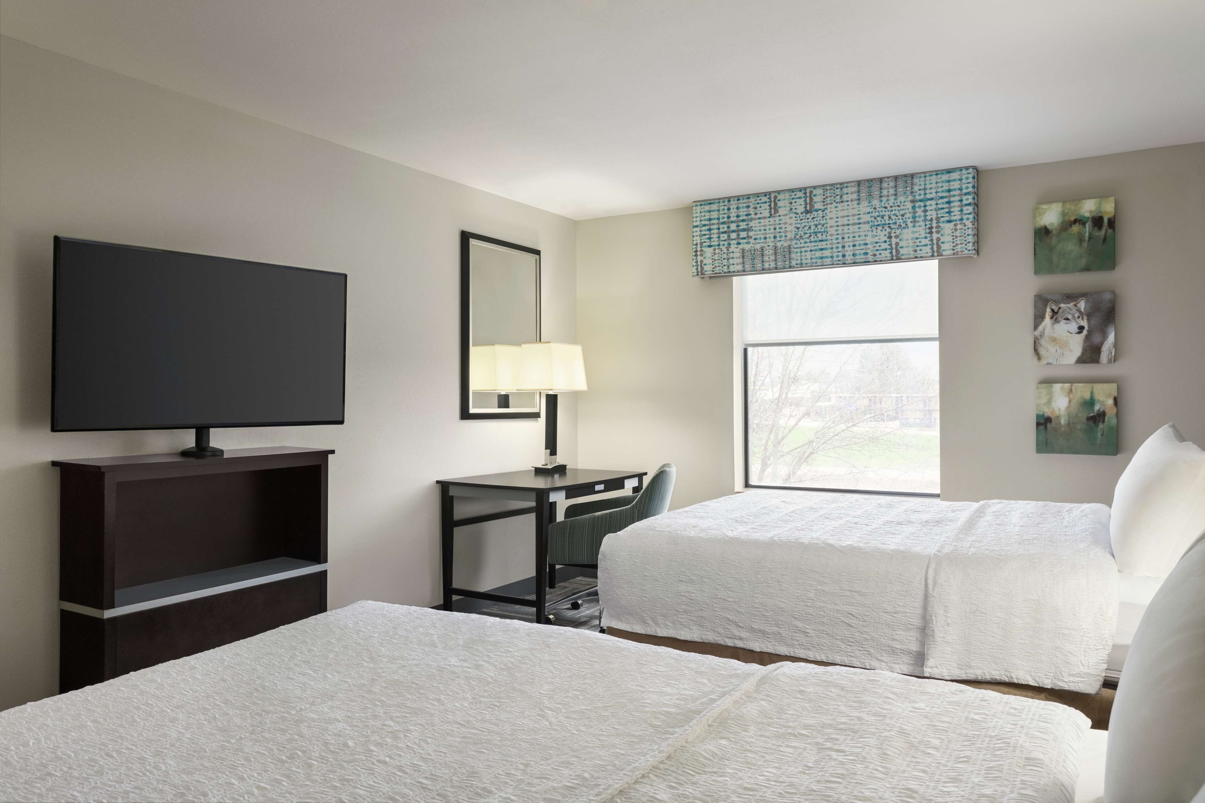 A hotel room with two beds, a desk, a TV, and artwork on the wall.