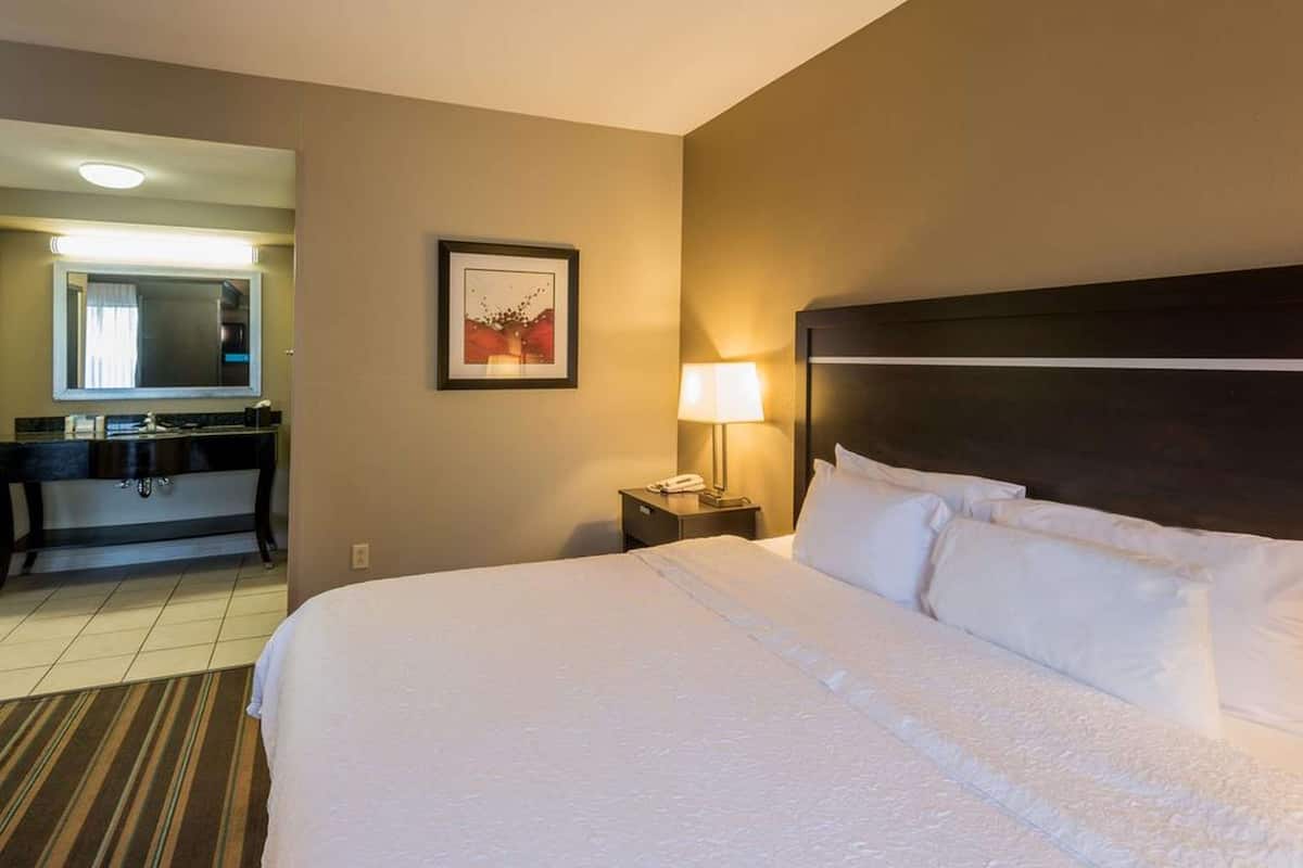 A hotel room with a large bed, a bedside table with a lamp, a framed picture on the wall, and a bathroom visible through an open door.
