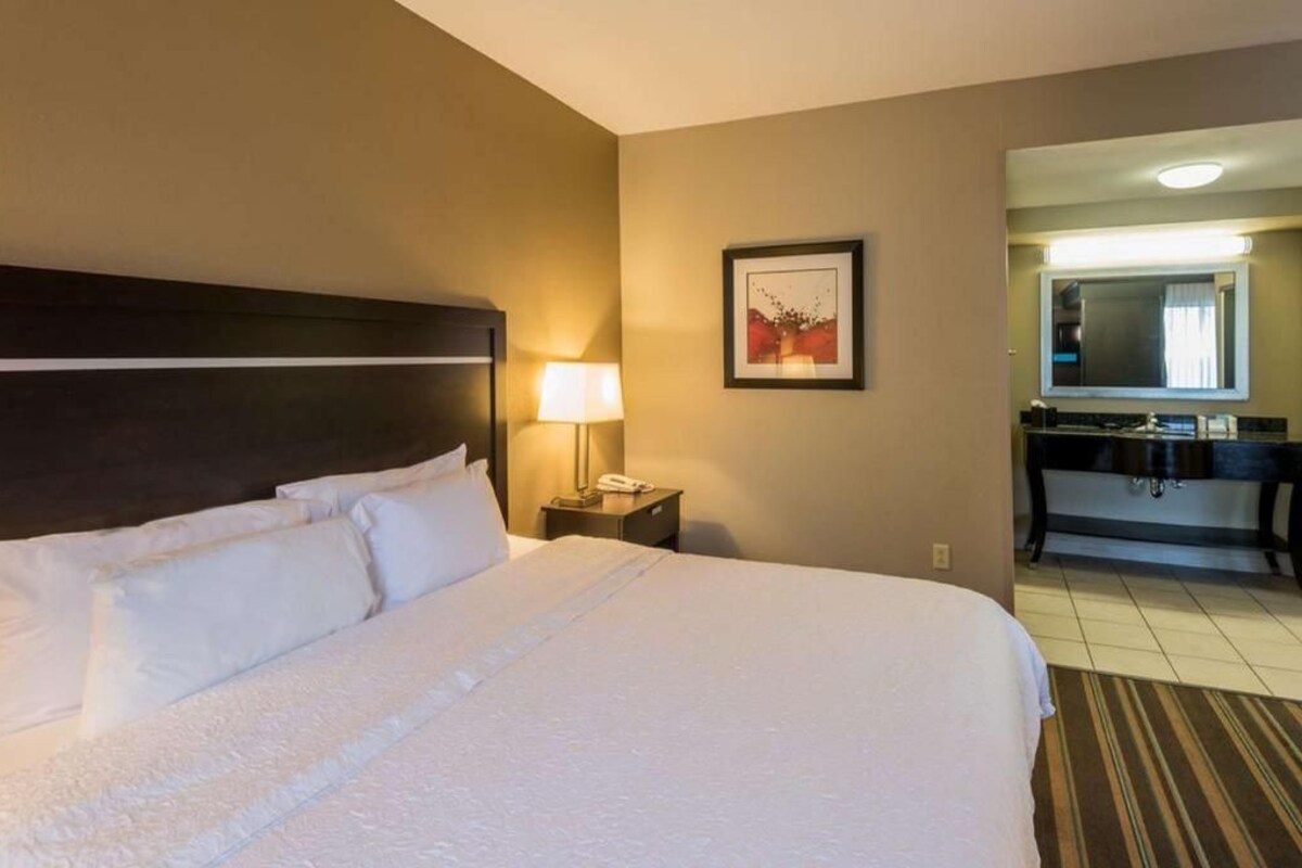 A hotel room with a large bed, a nightstand with a lamp, a framed picture on the wall, and a bathroom visible through an open door.