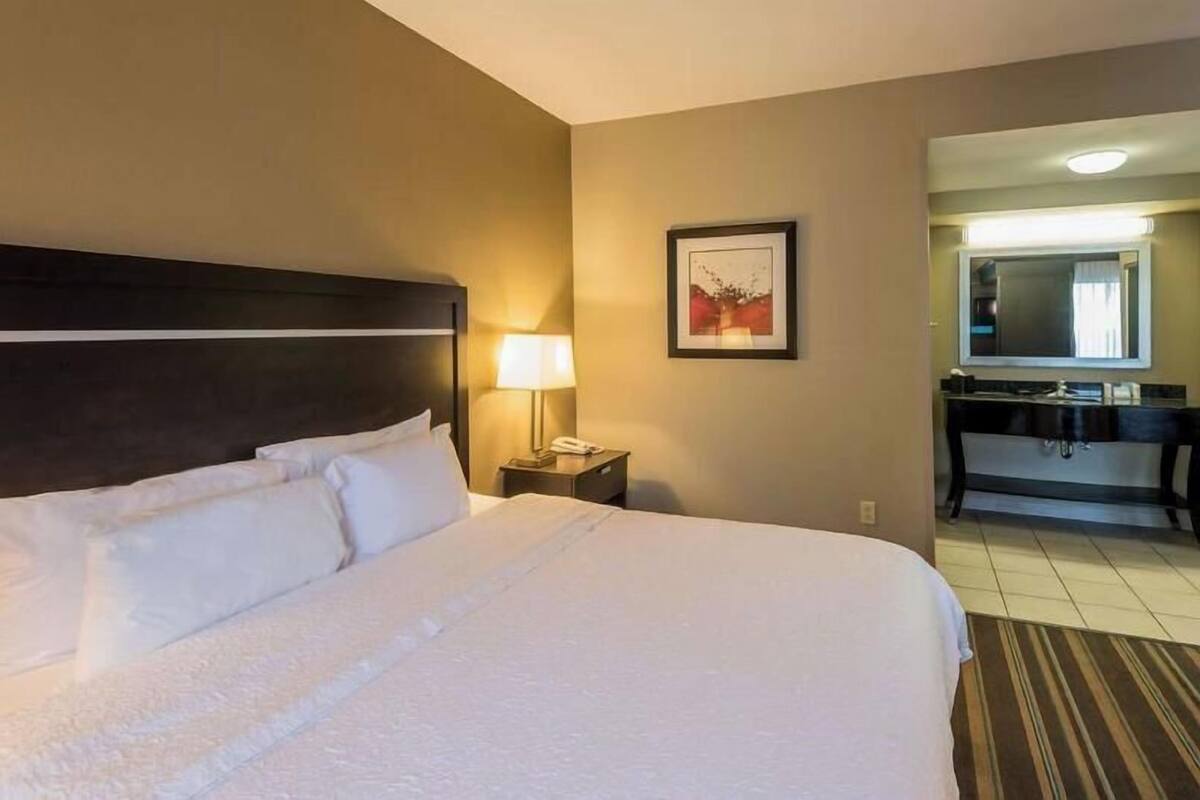 A hotel room with a large bed, a nightstand with a lamp, a framed picture on the wall, and a bathroom visible through an open door.