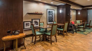 A hotel room with a wooden floor, a dining area with green chairs, a small table with a telephone, and framed pictures on the wall.