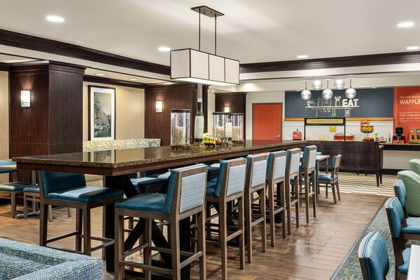 A modern hotel lobby with a central bar area, seating arrangements, and a breakfast counter.