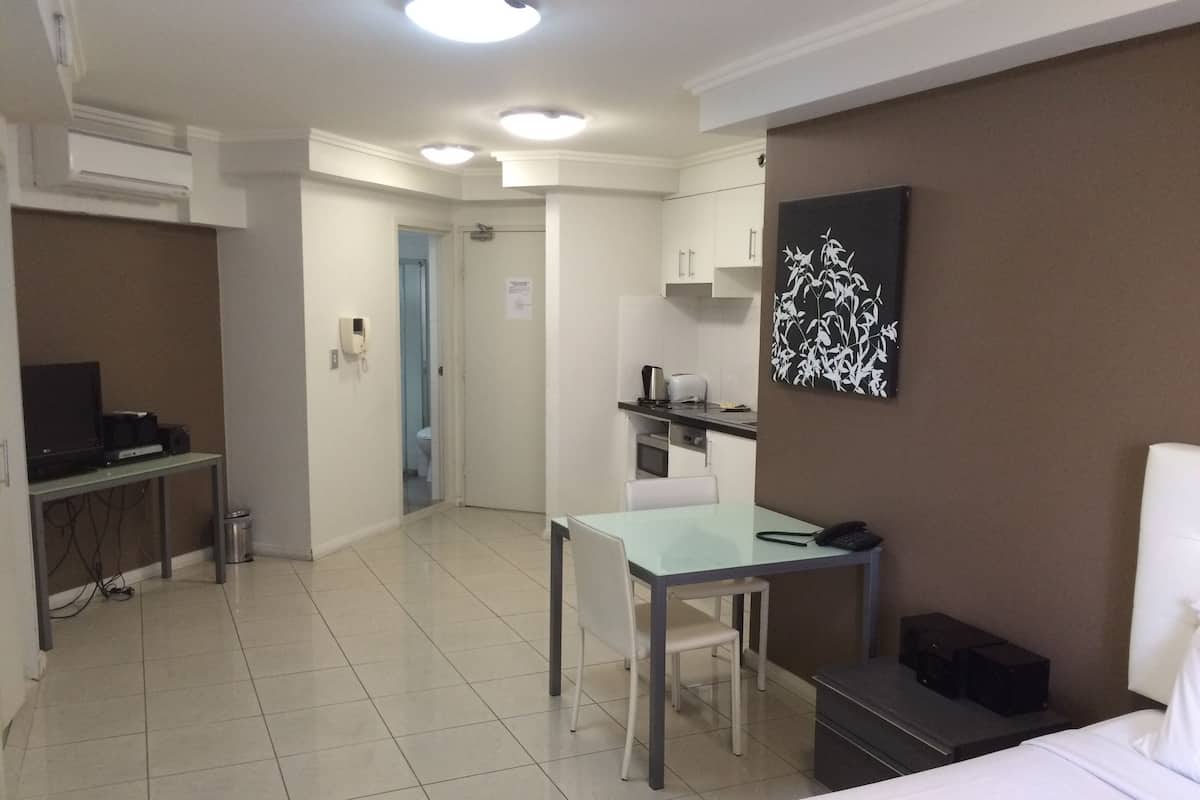 A hotel room with a bed, a desk with a chair, a small kitchenette, and a bathroom visible through an open door.