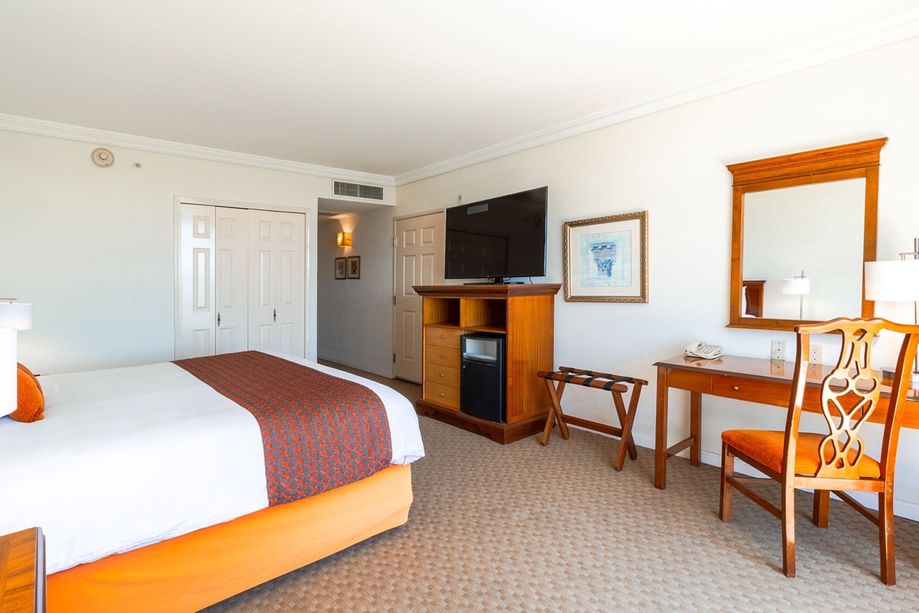 Quarto de hotel com cama, escrivaninha, cadeira, televisão e espelho.