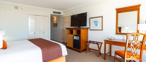Quarto de hotel com cama, escrivaninha, cadeira, televisão e espelho.