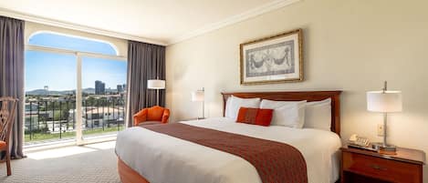 Habitación de hotel con una cama grande, una silla, una mesita de noche con lámpara y vistas de la ciudad a través de un ventanal amplio.