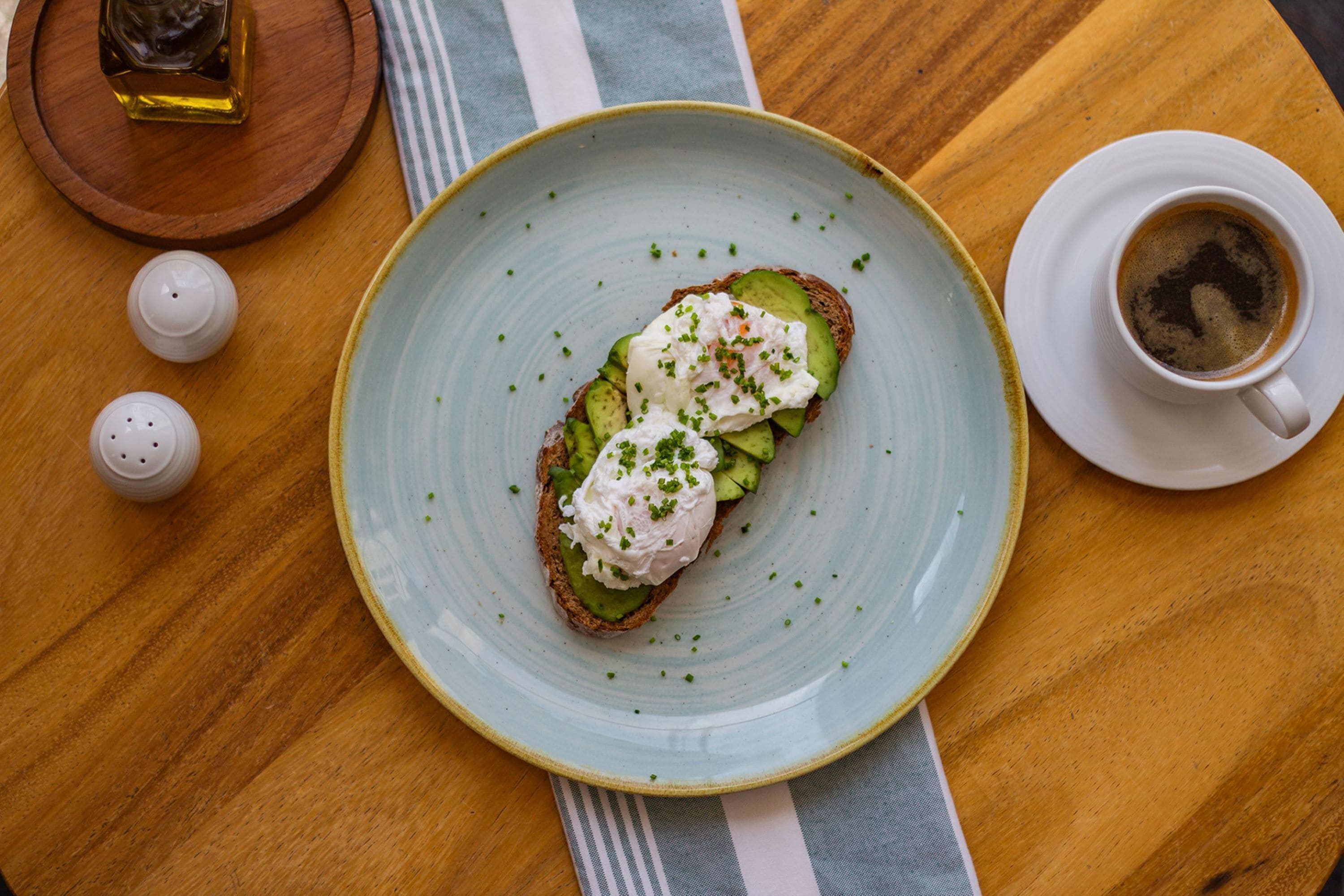 A plate with a sandwich topped with avocado and a poached egg, a cup of coffee, and salt and pepper shakers on a wooden table.