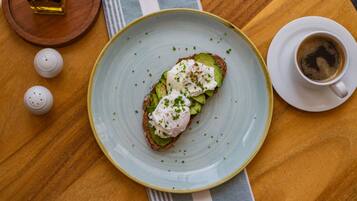 Een bord met een sandwich belegd met avocado en een gepocheerd ei, een kop koffie en zout- en peperstrooiers op een houten tafel.