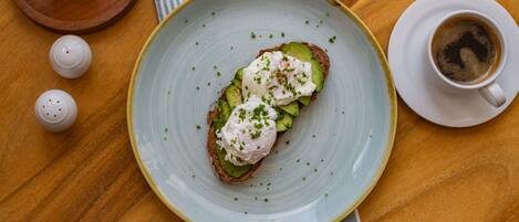 A plate with a sandwich topped with avocado and a poached egg, a cup of coffee, and salt and pepper shakers on a wooden table.